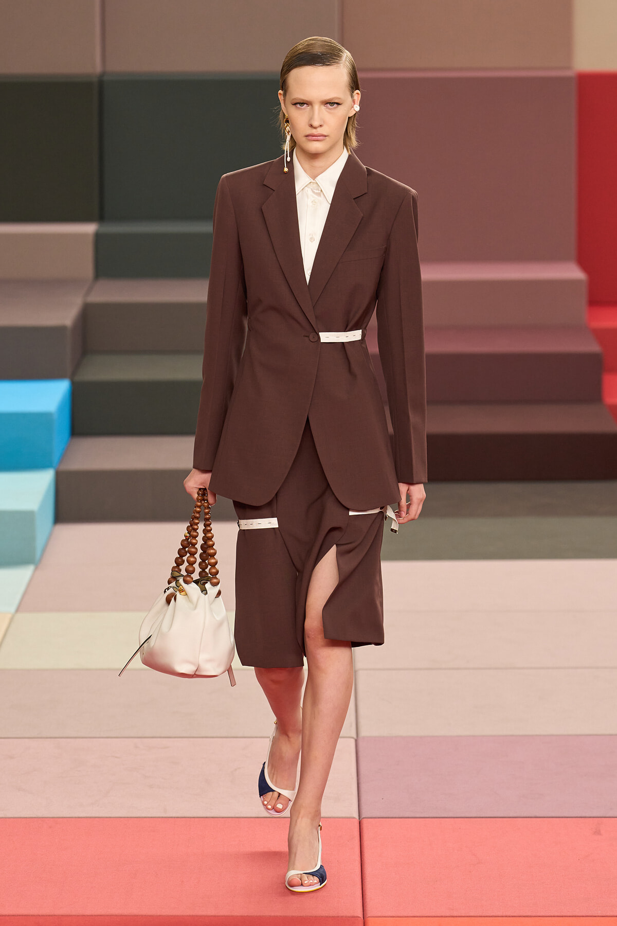 Model walking a runway in a brown suit with a white belt, white shirt, matching tailored blazer and split skirt; white bag with wooden bead handle hangs at her side.