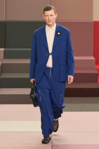 Male model walks a runway in a cobalt blue suit with a beige shirt, black belt, and black woven bag in hand.