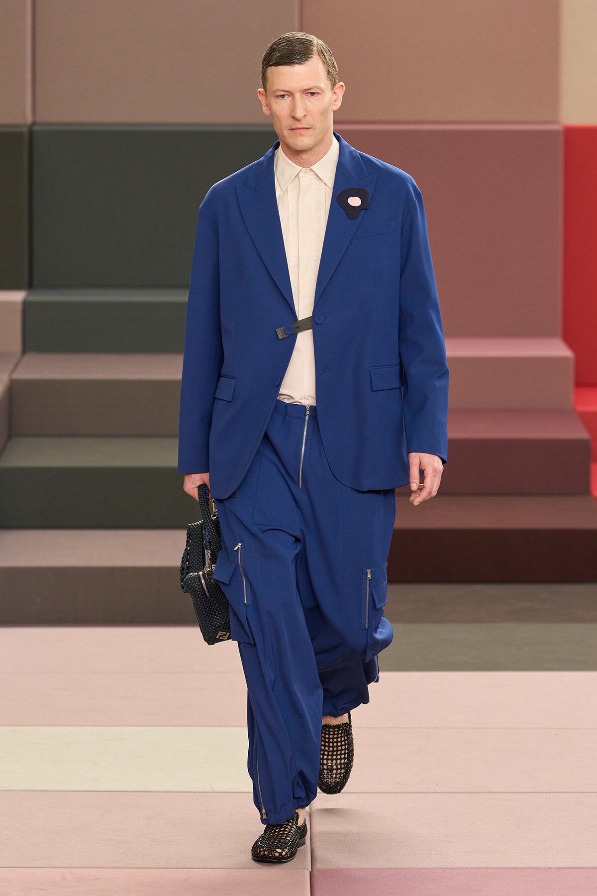 Male model walks a runway in a cobalt blue suit with a beige shirt, black belt, and black woven bag in hand.
