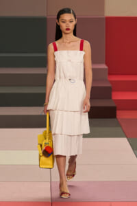 Model walking a fashion runway in a white pleated midi dress with red straps, carrying a yellow handbag with colorful pom-poms.