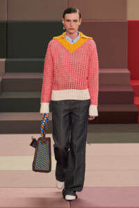 Runway model wearing a pink chunky knit sweater with a bright yellow V-neck collar over a blue shirt, paired with dark wide-leg jeans and white shoes, carrying a blue beaded handbag.