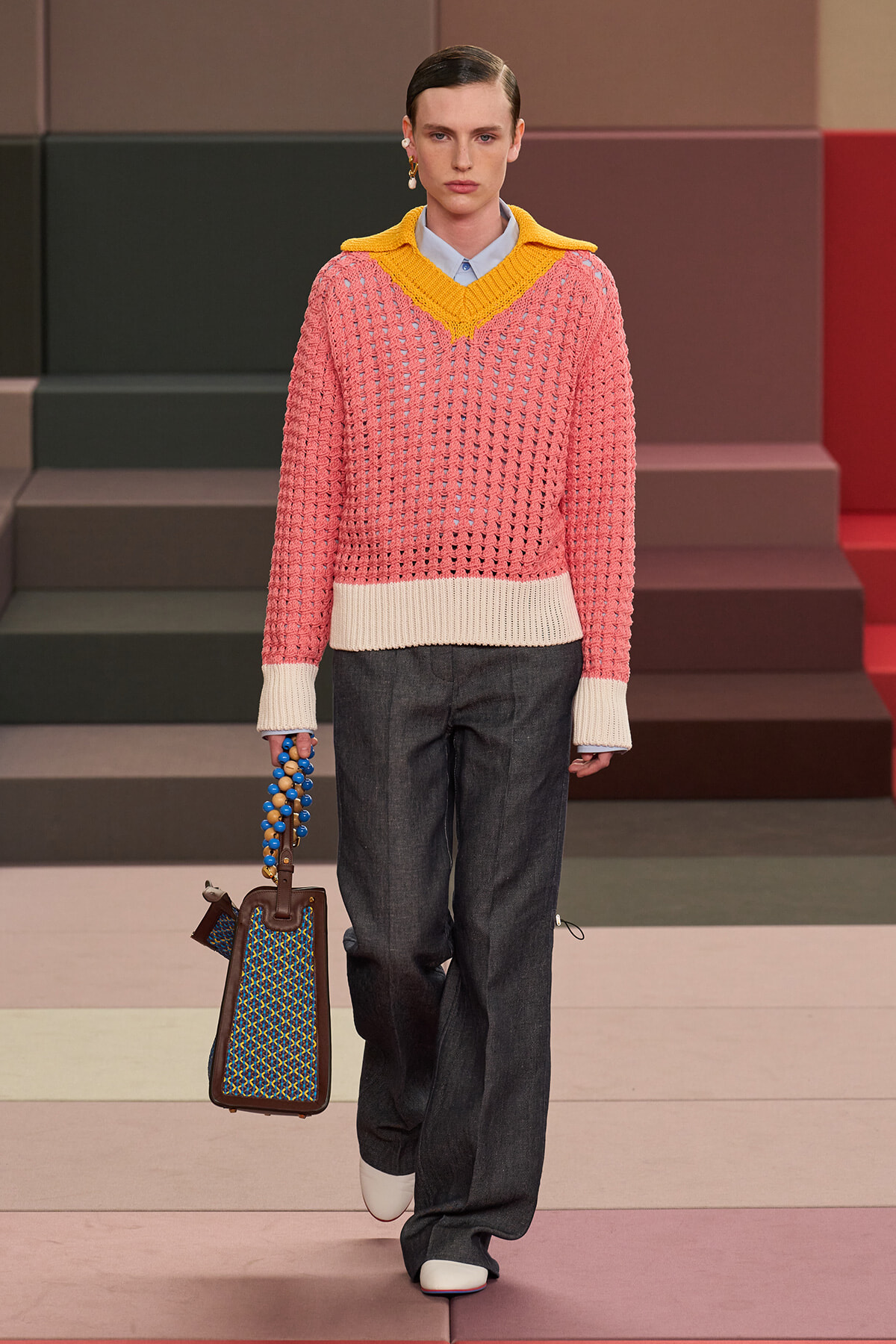 Runway model wearing a pink chunky knit sweater with a bright yellow V-neck collar over a blue shirt, paired with dark wide-leg jeans and white shoes, carrying a blue beaded handbag.