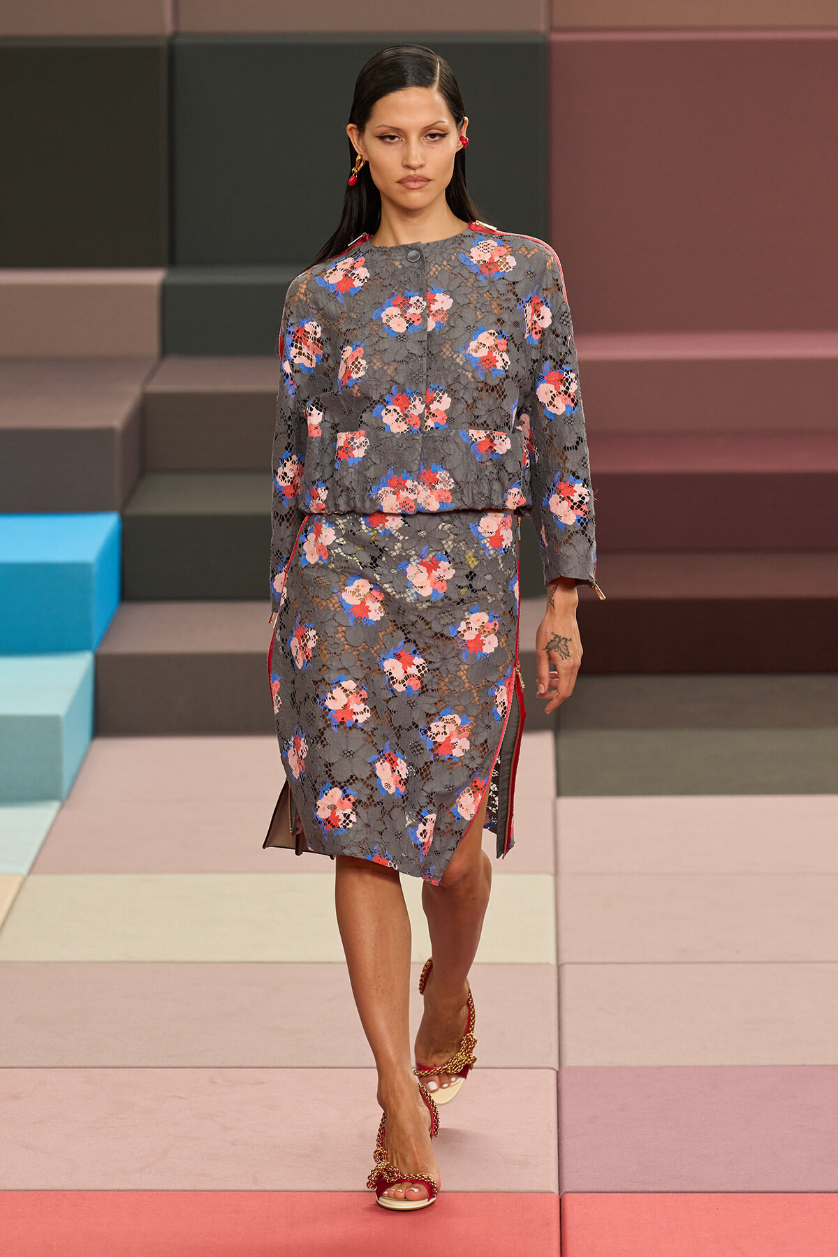 Model walking a runway in a gray floral lace dress with red, pink, and blue blooms, paired with red sandals.