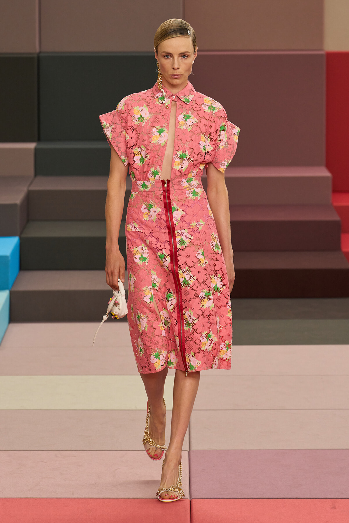 Runway model in a pink floral lace midi dress with a red center zipper, walking on a multicolor stepped runway.