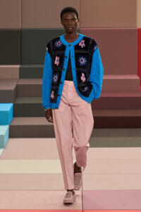 Runway model wearing a bright blue cardigan with pink-purple floral patches over a black top, paired with pale pink trousers and pink woven shoes.