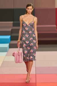 Model walks a fashion runway in a gray floral lace slip dress with slender straps, carrying a pink faux‑fur handbag against a multicolored stepped backdrop.