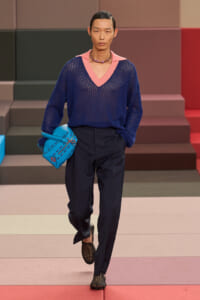 Male model on a runway wearing a navy blue mesh V-neck sweater with pink trim, dark trousers, black mesh shoes, and a bright turquoise handbag.