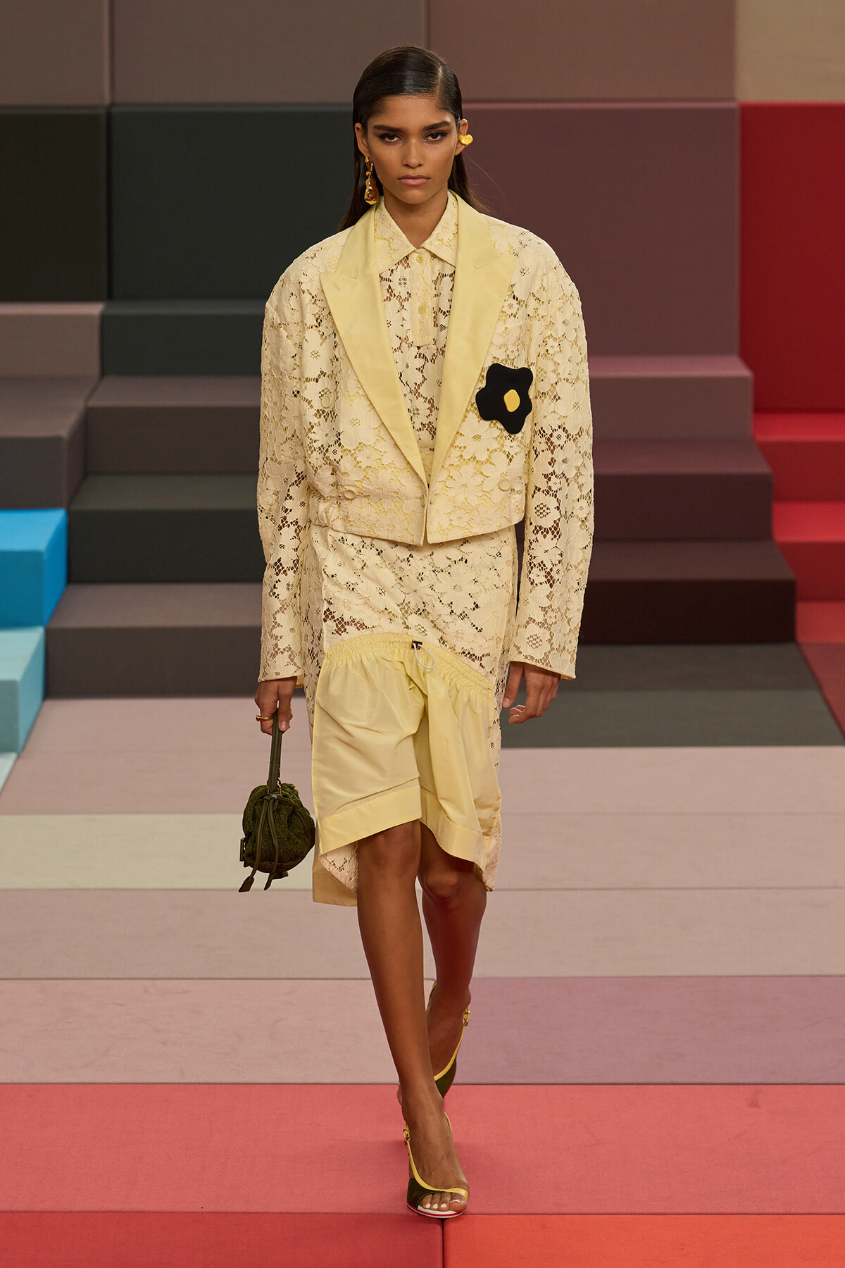 Model walking a runway in a pale yellow lace suit with cream lapels and a black flower brooch on the blazer, carrying a small green bag and wearing yellow sandals.