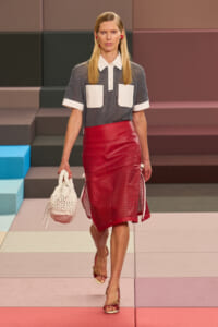 Runway model wearing a gray knit polo with white pockets and collar, a red perforated midi skirt, red sandals, and a white crocheted bag.