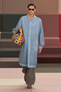 Model on a runway wearing a light blue perforated coat, gray wide-leg pants, brown woven sandals, sunglasses, and a colorful checkered tote bag.