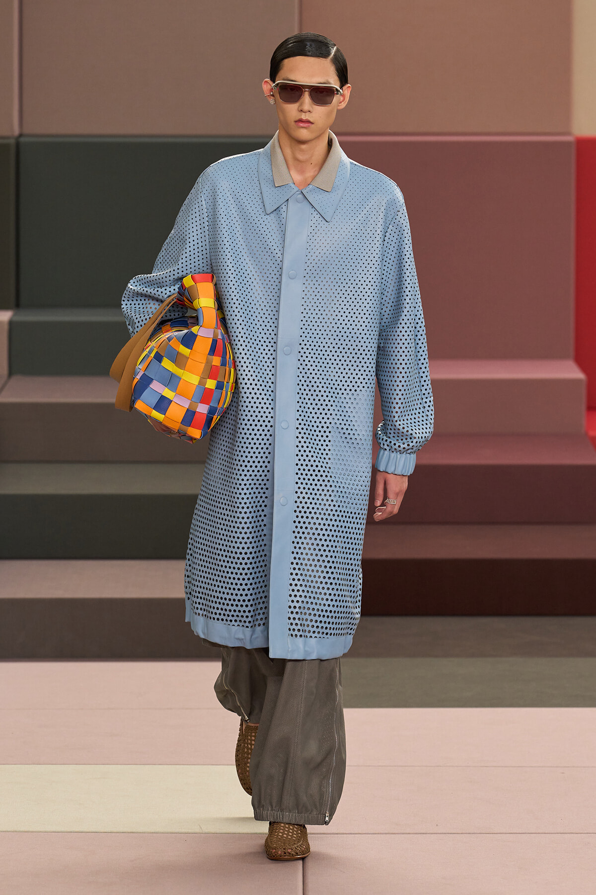 Model on a runway wearing a light blue perforated coat, gray wide-leg pants, brown woven sandals, sunglasses, and a colorful checkered tote bag.