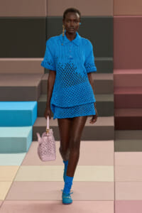 Female model walks a runway in a blue crochet polo and matching mini skirt, carrying a pale pink woven handbag.