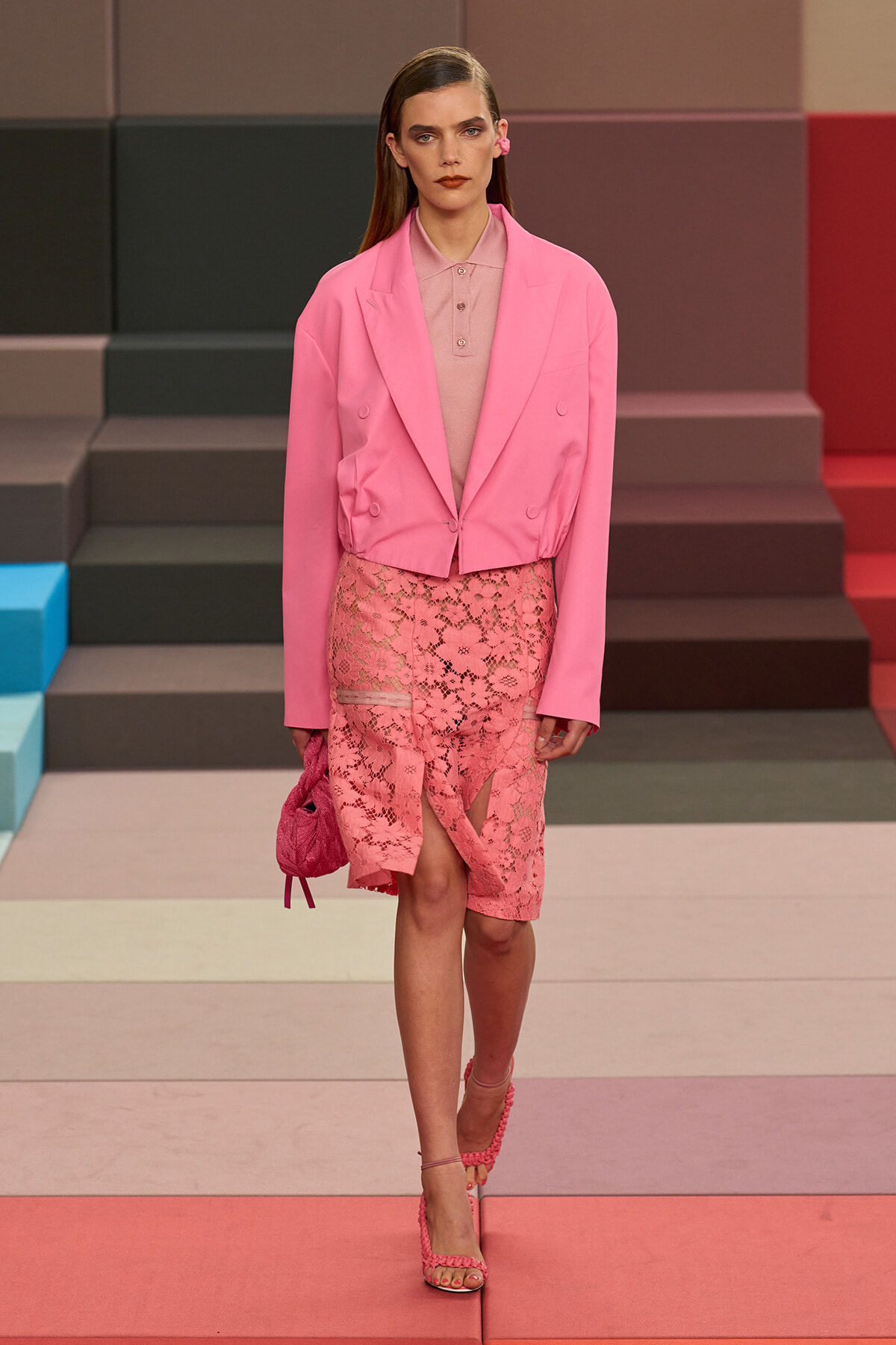 Model on a runway wearing a bright pink double‑breasted blazer over a pink polo, with a pink lace skirt and matching handbag.