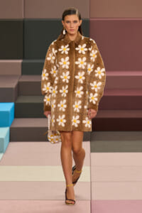 Model wearing a knee‑length brown coat with large white daisy patterns on a pastel runway.