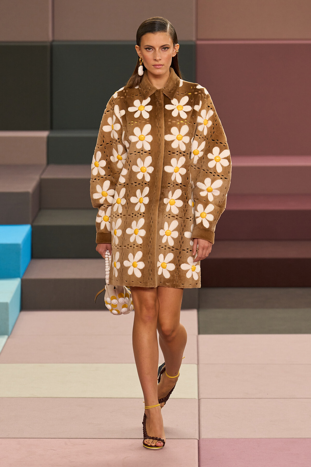 Model wearing a knee‑length brown coat with large white daisy patterns on a pastel runway.