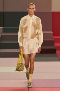 Model walking runway in cream abstract-pattern shirt and matching shorts, holding a yellow net bag with gray interior.