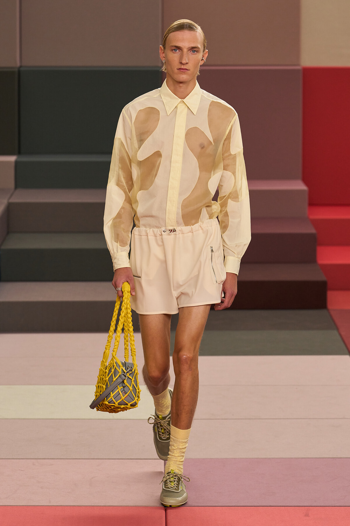 Model walking runway in cream abstract-pattern shirt and matching shorts, holding a yellow net bag with gray interior.
