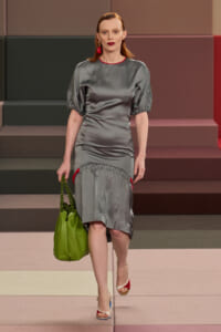 Model walking a fashion runway in a gray satin dress with puff sleeves, red trim and red earrings, carrying a bright green handbag.