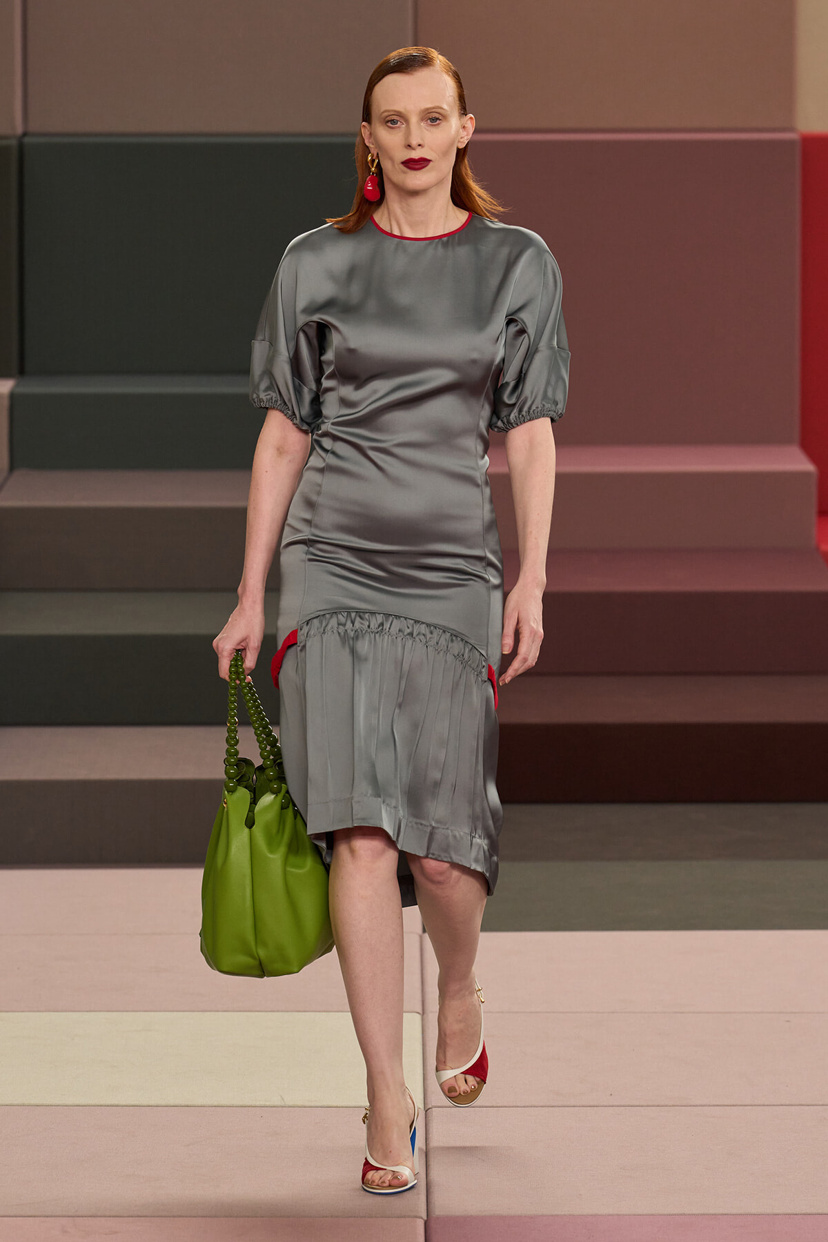 Model walking a fashion runway in a gray satin dress with puff sleeves, red trim and red earrings, carrying a bright green handbag.