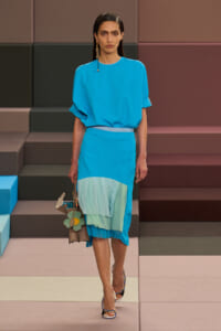 Runway model in a bright blue dress with a layered mint skirt, carrying a brown handbag with flower charms and wearing statement earrings on a stepped, colorful set.