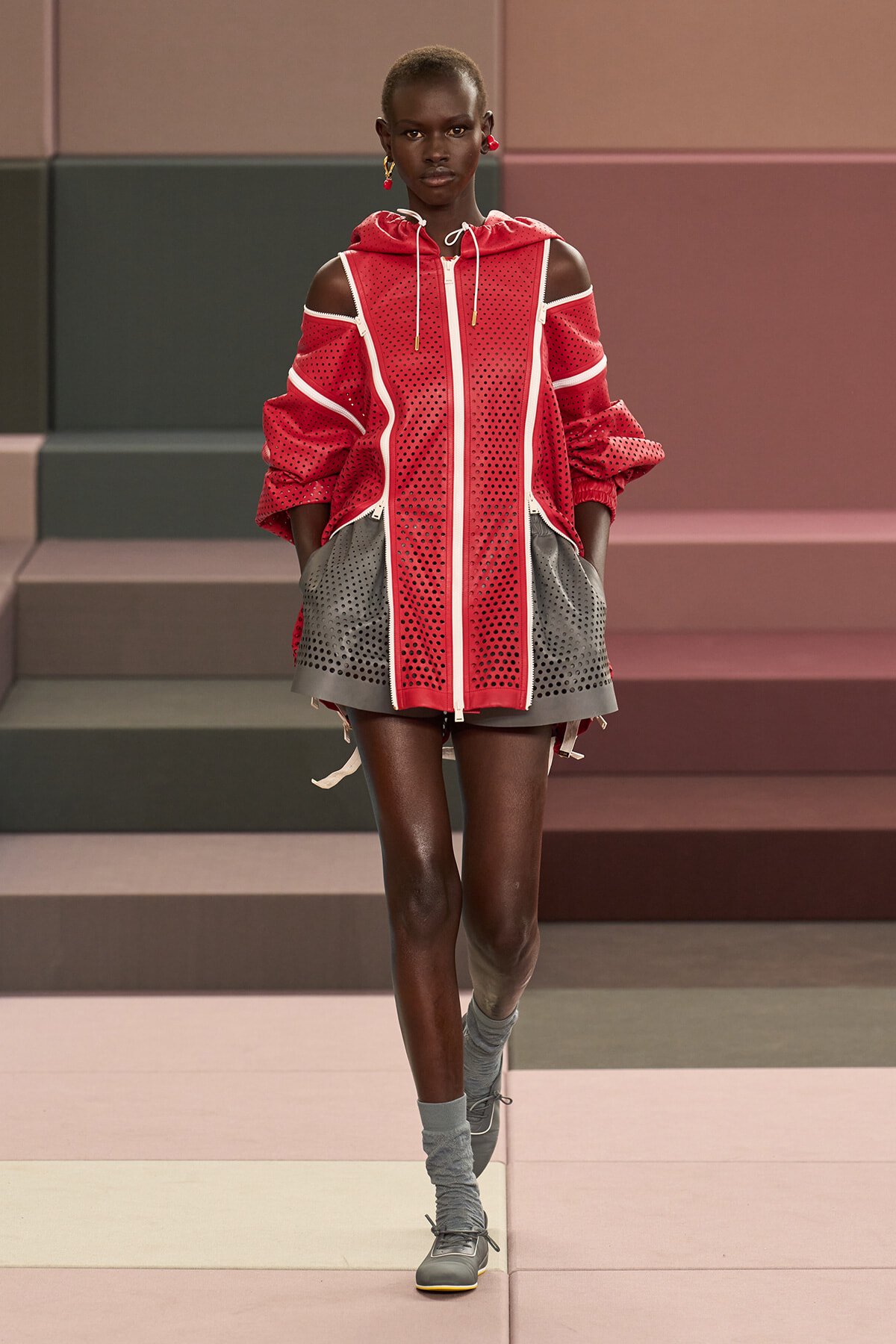 Runway model in a red perforated hooded dress with white piping and gray perforated pockets, paired with gray socks and sneakers on a pastel-toned catwalk.