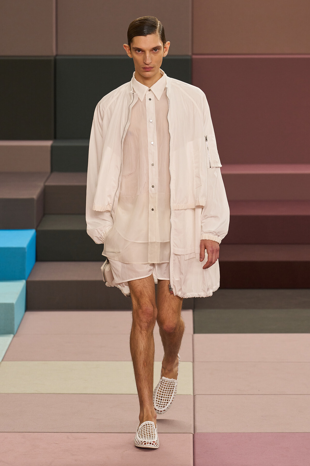 Male model walking a fashion runway in an all-white layered outfit: sheer shirt, light jacket, shorts, and white lattice loafers/Shoes.