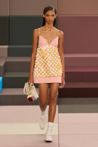 Model walking a runway in a pink slip dress with white and yellow circular appliqués, carrying a cream handbag with a multicolored pom-pom.