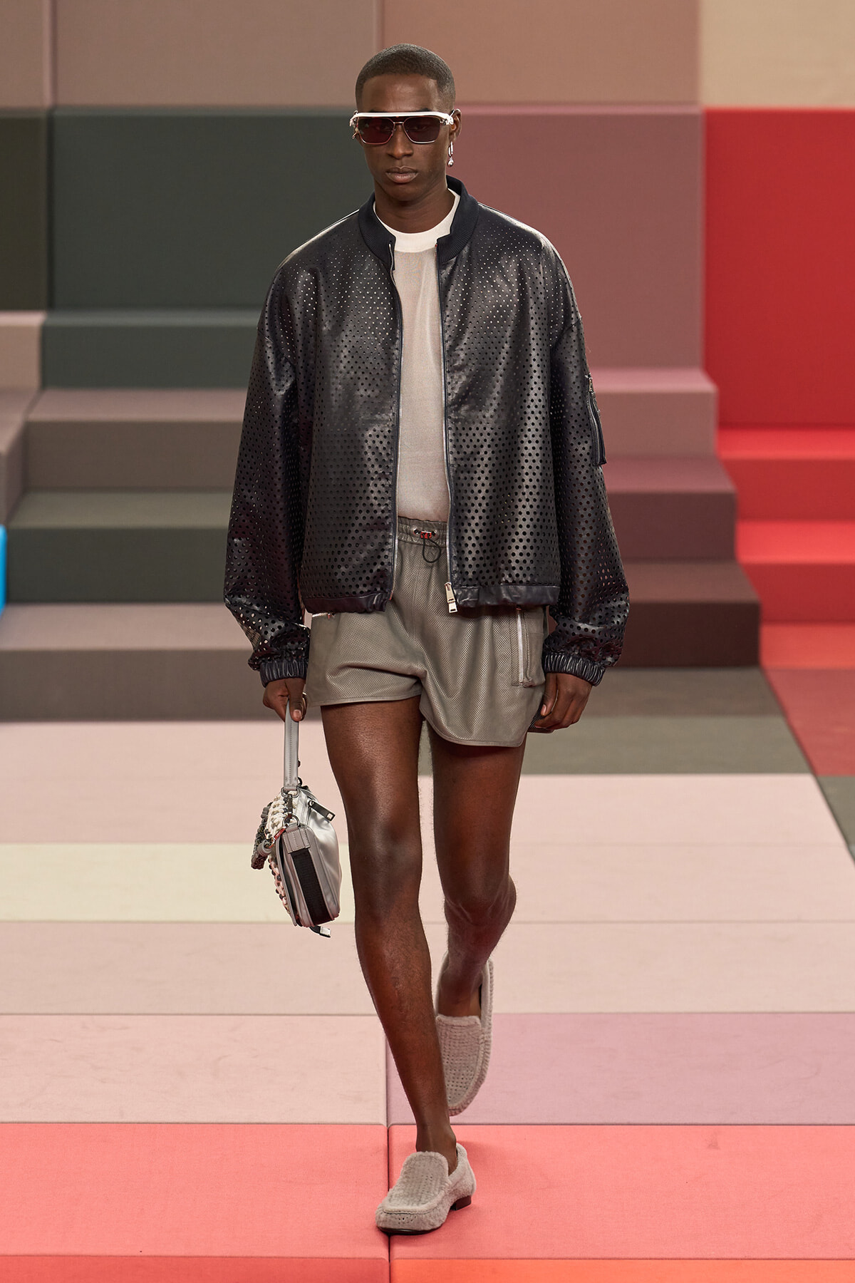 Male model walks a fashion show wearing a perforated black bomber jacket, gray shorts, white shirt, white sunglasses, and carries a small gray handbag on the runway.