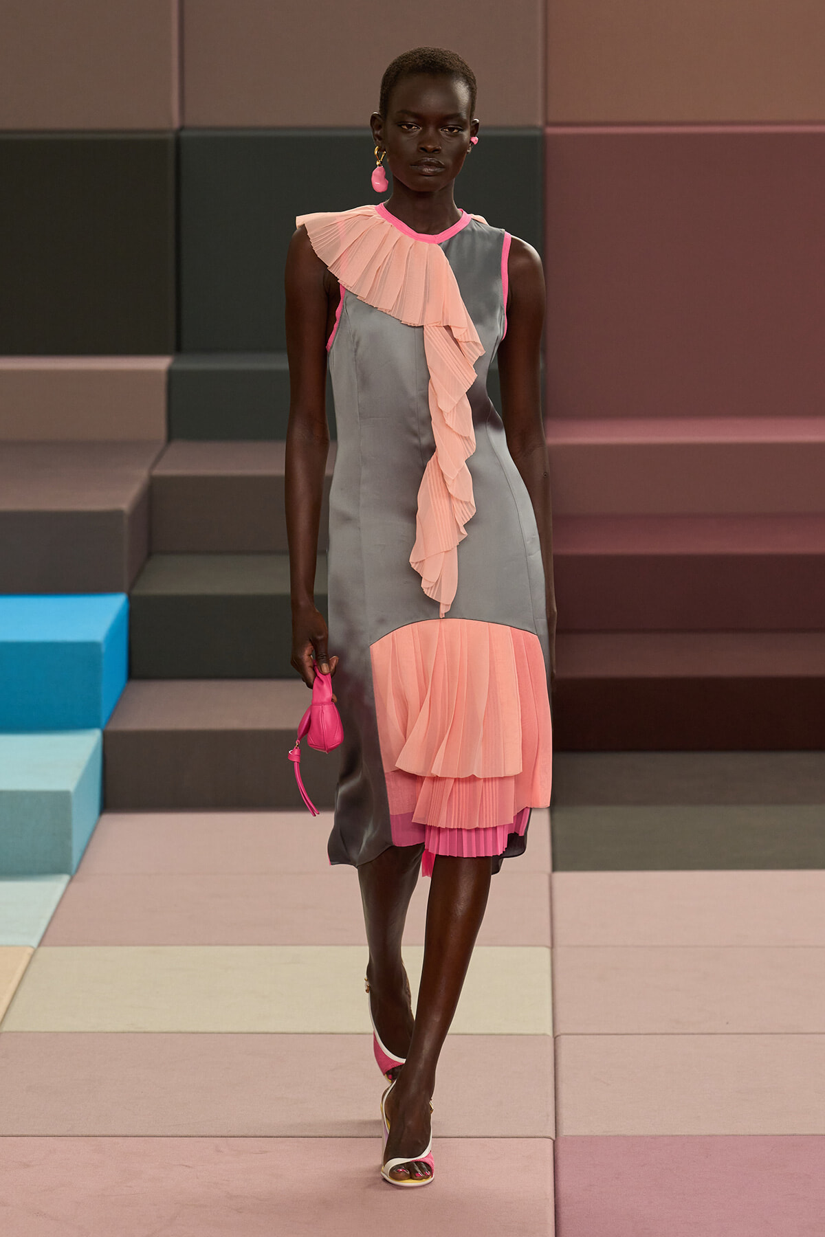 Model walking a fashion runway in a gray sleeveless midi dress with cascading peach-pink pleated ruffles from shoulder to hem, paired with pink accessories.