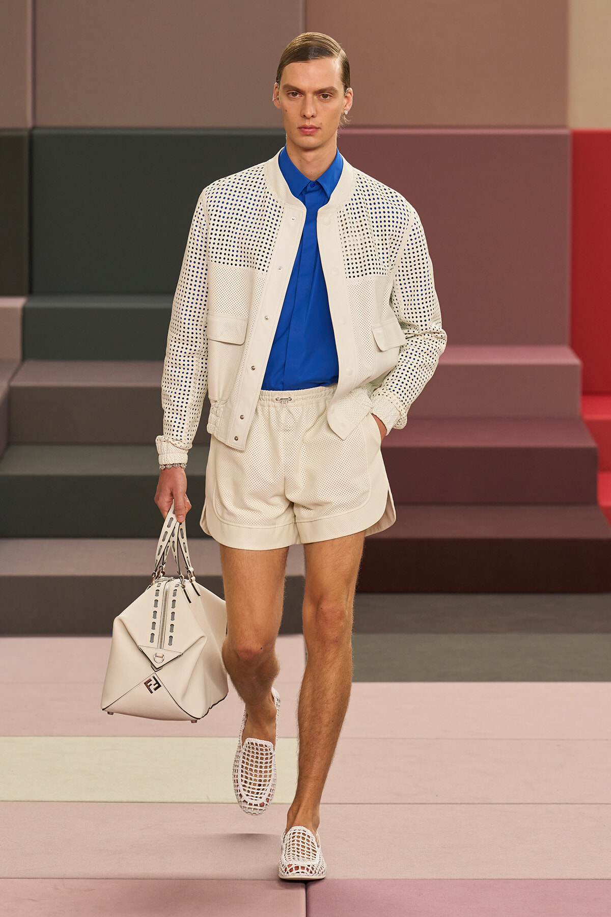 Male model walking runway in a royal blue shirt with a white perforated jacket and matching shorts, carrying a white structured bag with perforated sandals.