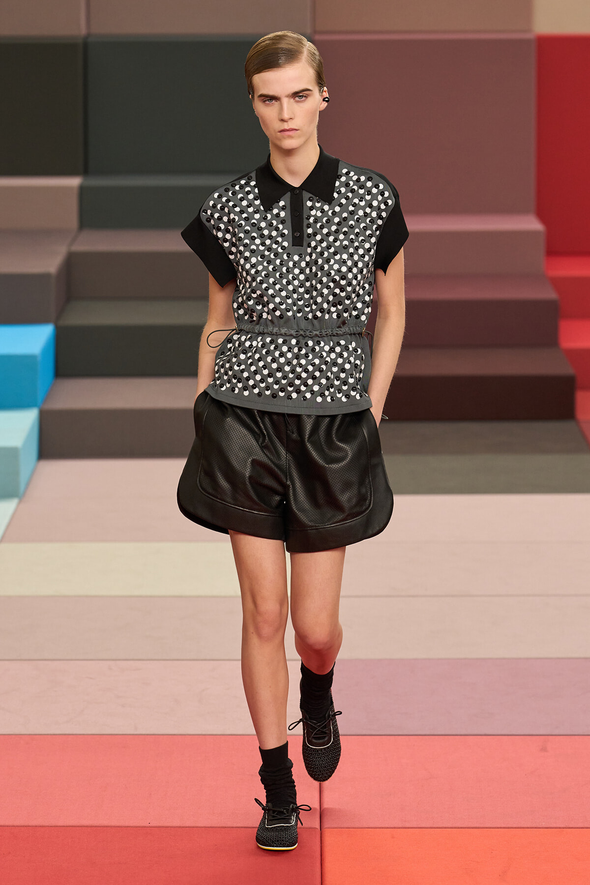 Model walking a fashion runway in a black-and-white polka-dot top with black shorts and belt, against a multicolored stepped backdrop.