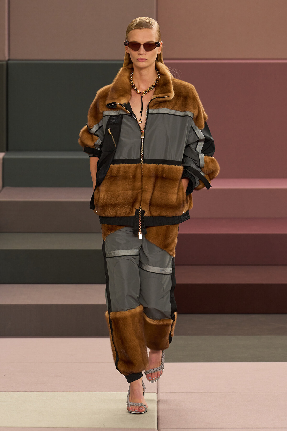 Model on a runway wearing a brown fur-trimmed gray jacket with matching pants and sunglasses.