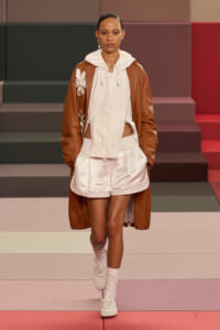 Female model walks down a fashion runway wearing a white hoodie, white satin shorts, and a brown long coat with a floral appliqué on the sleeve; colorful panel backdrop.