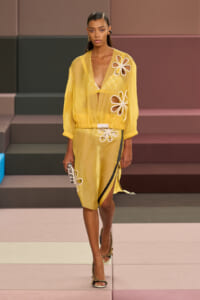 Model wearing a sheer yellow cardigan and matching skirt with large white flower appliqués on a pastel runway stage in heels, holding a small patterned clutch.