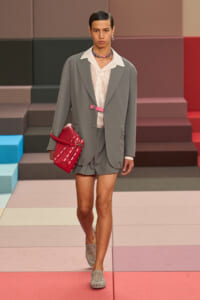 Model walks the runway in a gray blazer with matching shorts, white shirt, pink belt, and a red handbag.