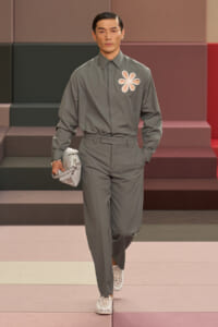 Male model in a gray matching shirt and trousers with a white floral emblem on the chest, walking a fashion runway and holding a light gray clutch.