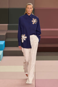 Model walking a runway in a navy button-down shirt with white flower appliqués and cream wide-leg trousers.