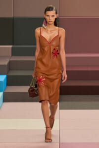 Model on a runway wearing a caramel leather slip dress with red trim and red floral cutouts, carrying a brown beaded-handled bag and wearing red sandals.