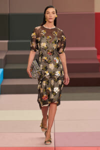 Model walking the runway in a sheer black floral midi dress with yellow and white blooms, carrying a beaded clutch.
