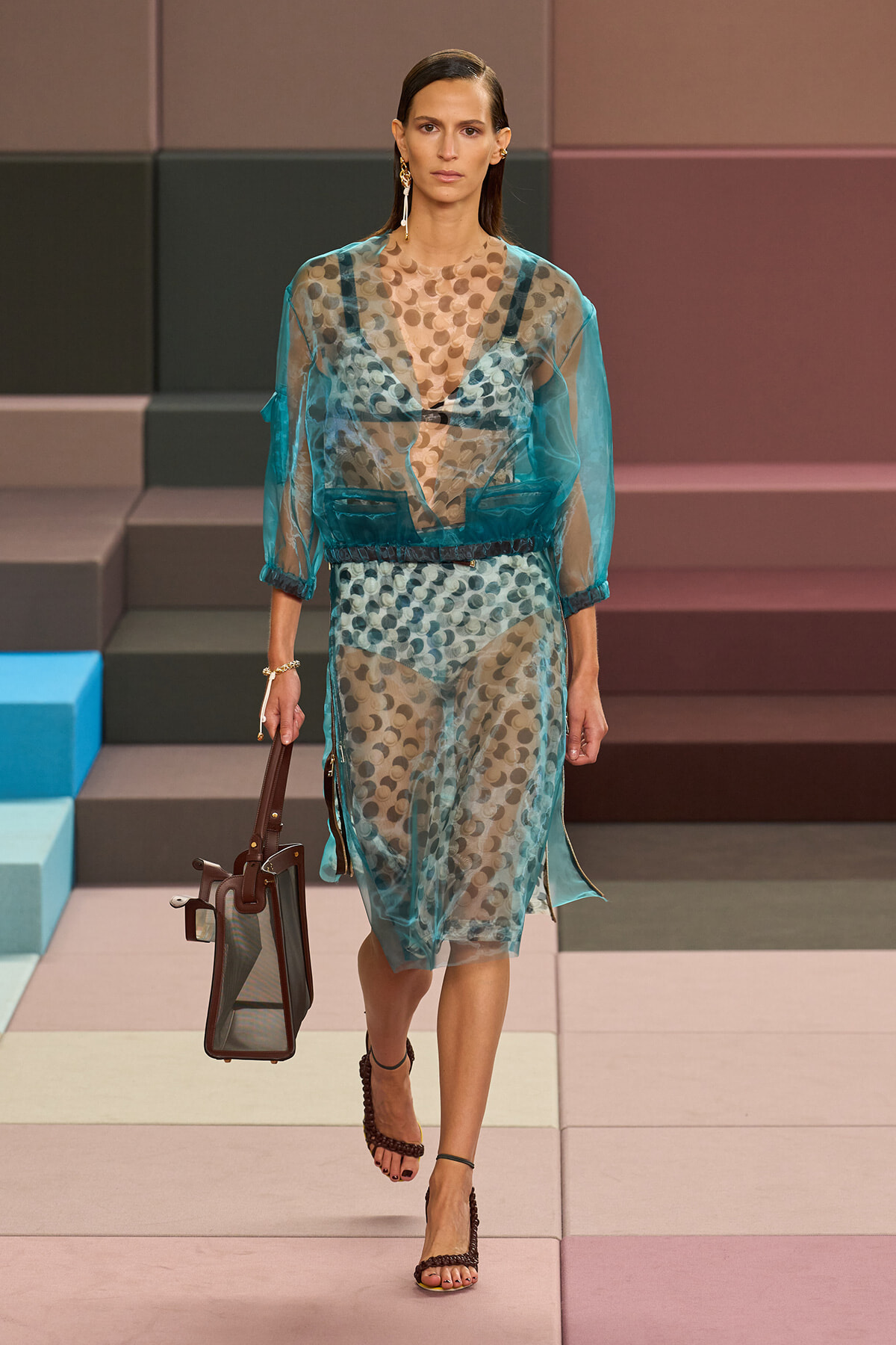Model walks the runway wearing a sheer teal polka‑dot dress over a matching underlayer, carrying a brown structured tote and wearing brown strappy sandals in a fashion show setting.