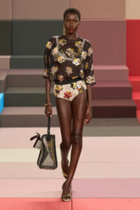 Model on a runway wearing a sheer black floral blouse with matching high-waisted floral shorts, carrying a clear structured handbag.