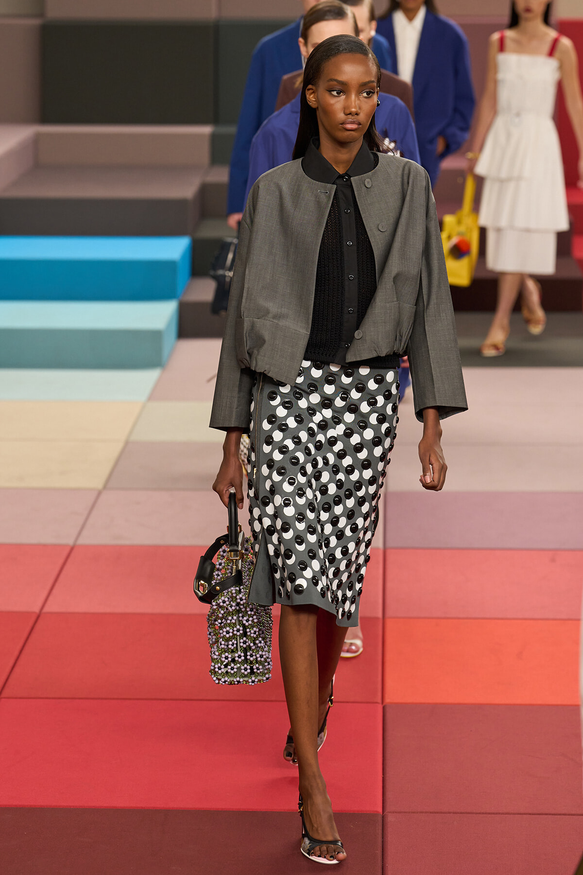Model walking a fashion runway in a gray cropped blazer, black blouse, and a black-and-white polka-dot midi skirt, carrying a beaded handbag visible at her side.