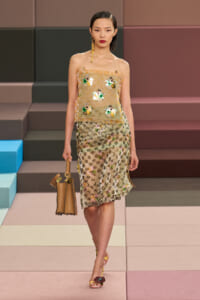 Model walks a fashion runway in a sheer, strapless beige floral top with appliqué flowers and a matching translucent skirt, carrying a tan handbag.