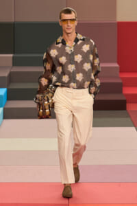 Male model walking a runway in a sheer brown floral shirt, cream trousers, yellow sunglasses, woven loafers, and a checkered bag under his arm