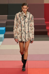 Model walking a fashion runway in a cream floral-printed oversized jacket with black piping and zippers, on a colorful tiled backdrop.