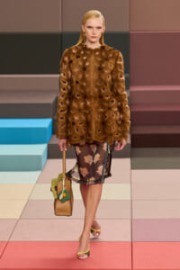 Model in a brown fur coat with cutout details over a floral sheer dress walks a pastel-tiled runway, carrying a tan handbag with green embellishments and wearing yellow-green sandals.