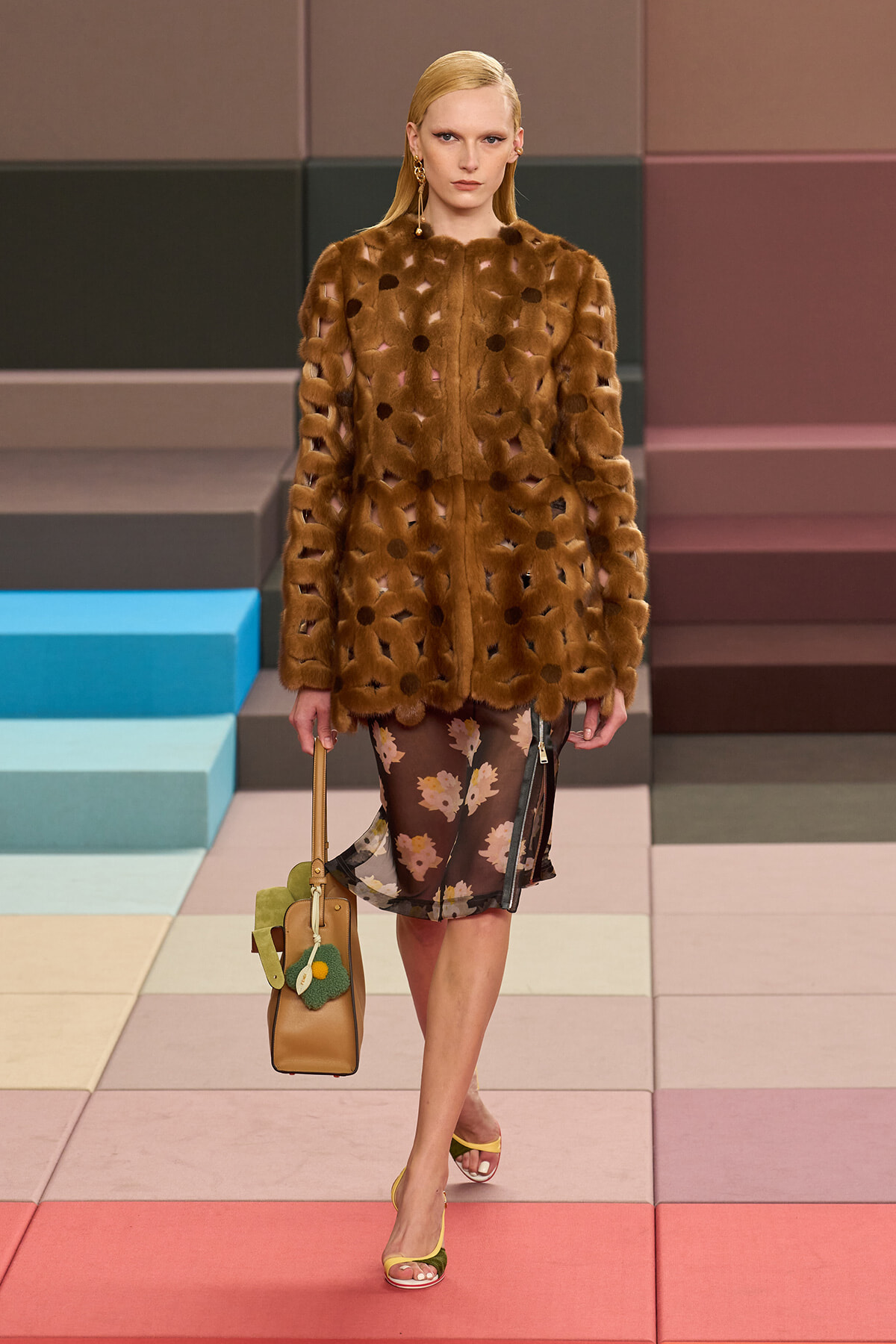 Model in a brown fur coat with cutout details over a floral sheer dress walks a pastel-tiled runway, carrying a tan handbag with green embellishments and wearing yellow-green sandals.