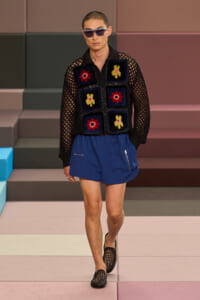 Male model walking a runway in a black mesh knit top with colorful square patches and blue shorts, wearing sunglasses.