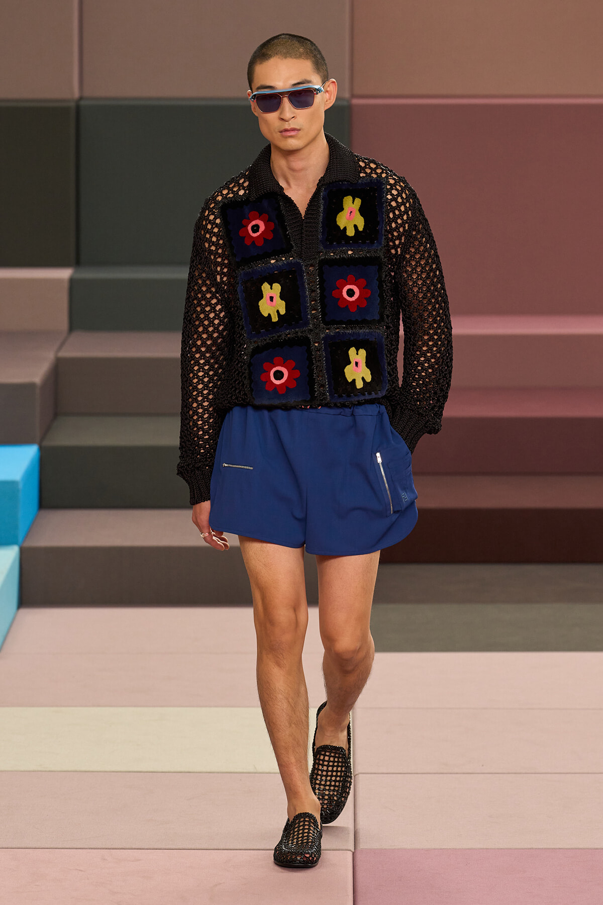 Male model walking a runway in a black mesh knit top with colorful square patches and blue shorts, wearing sunglasses.