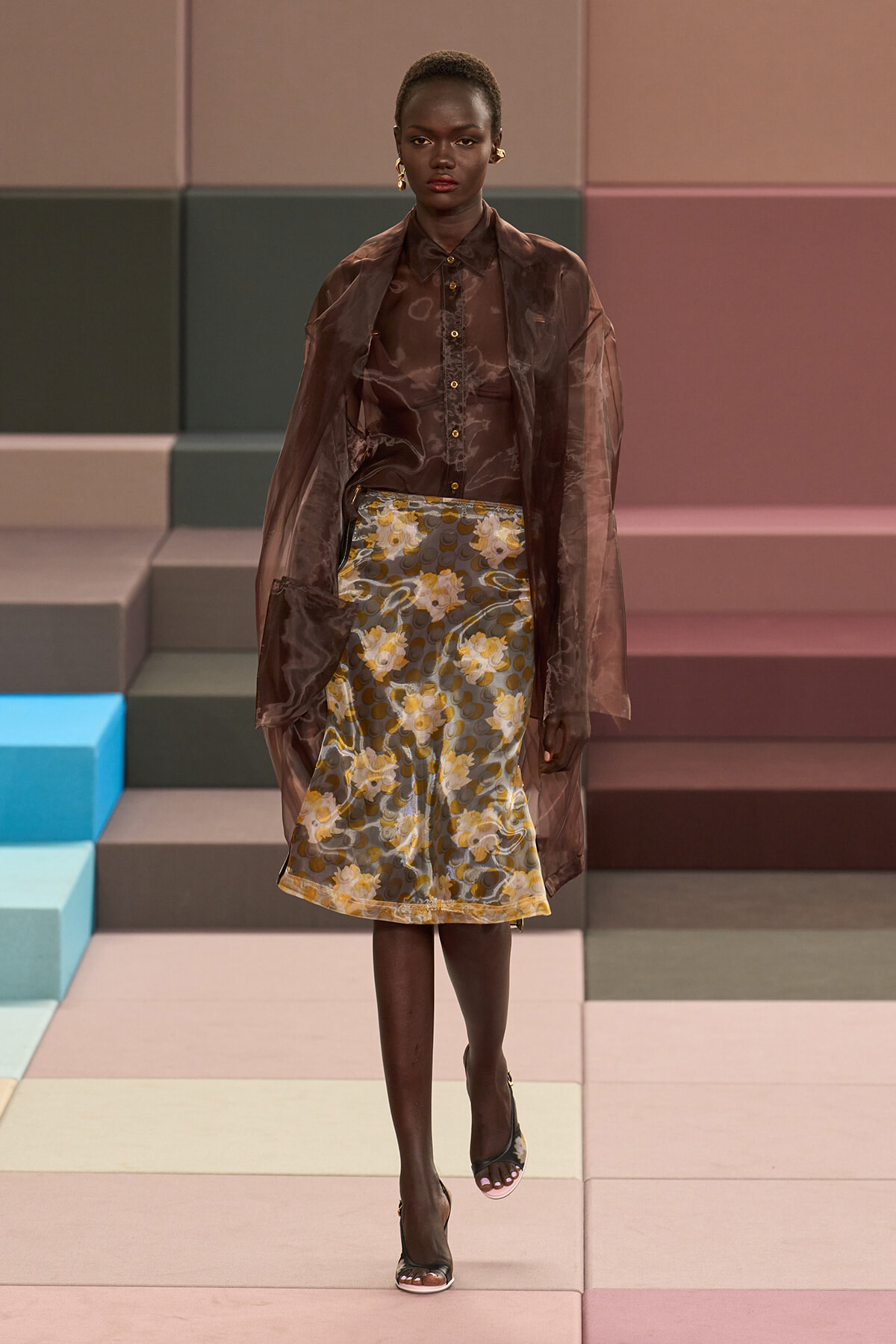 Model wearing a sheer brown blouse and matching coat over a floral A-line skirt on a pastel-toned runway edges?